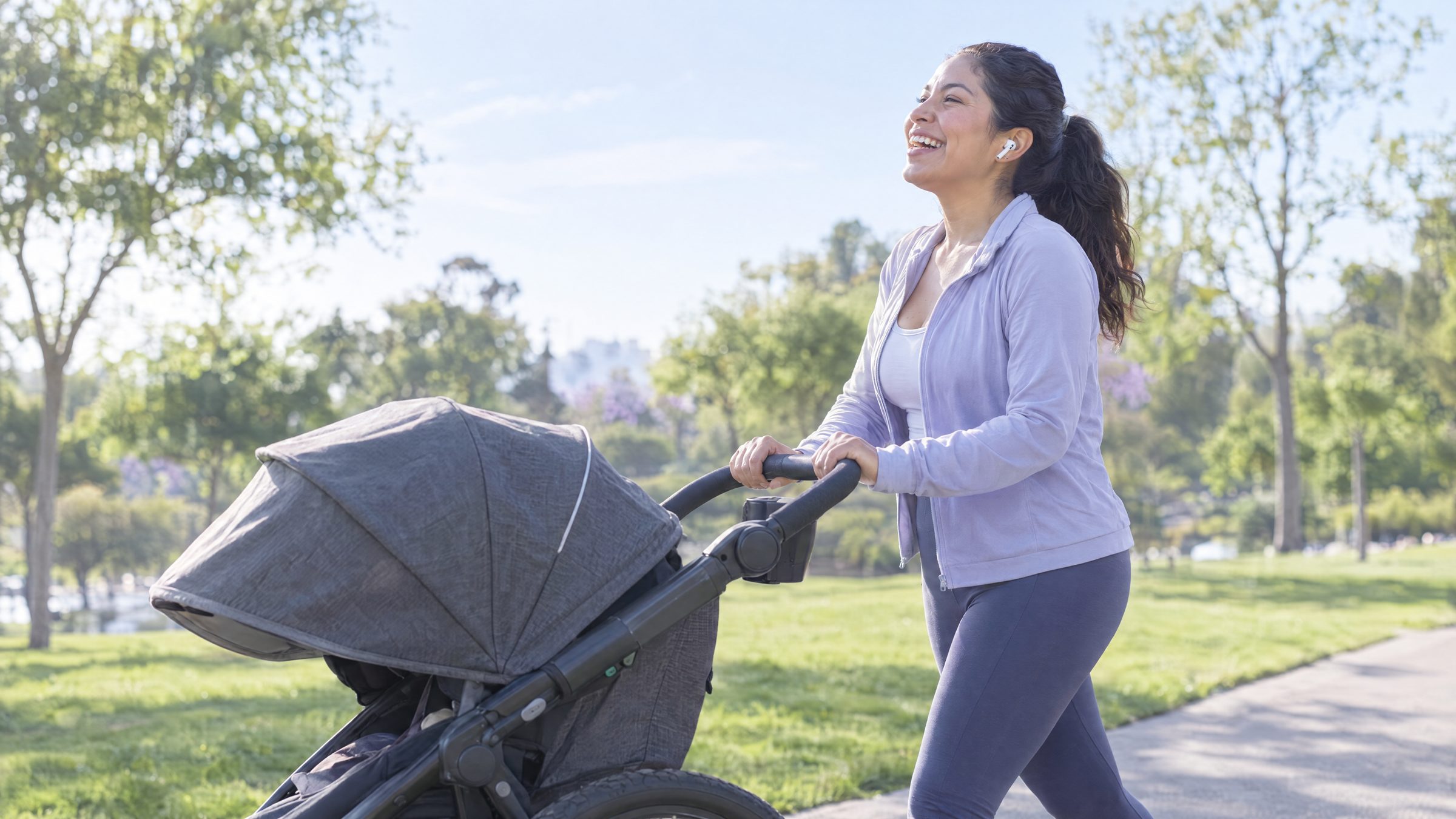 Woman laughing through earbuds as she pushes a jogging stroller along a park path